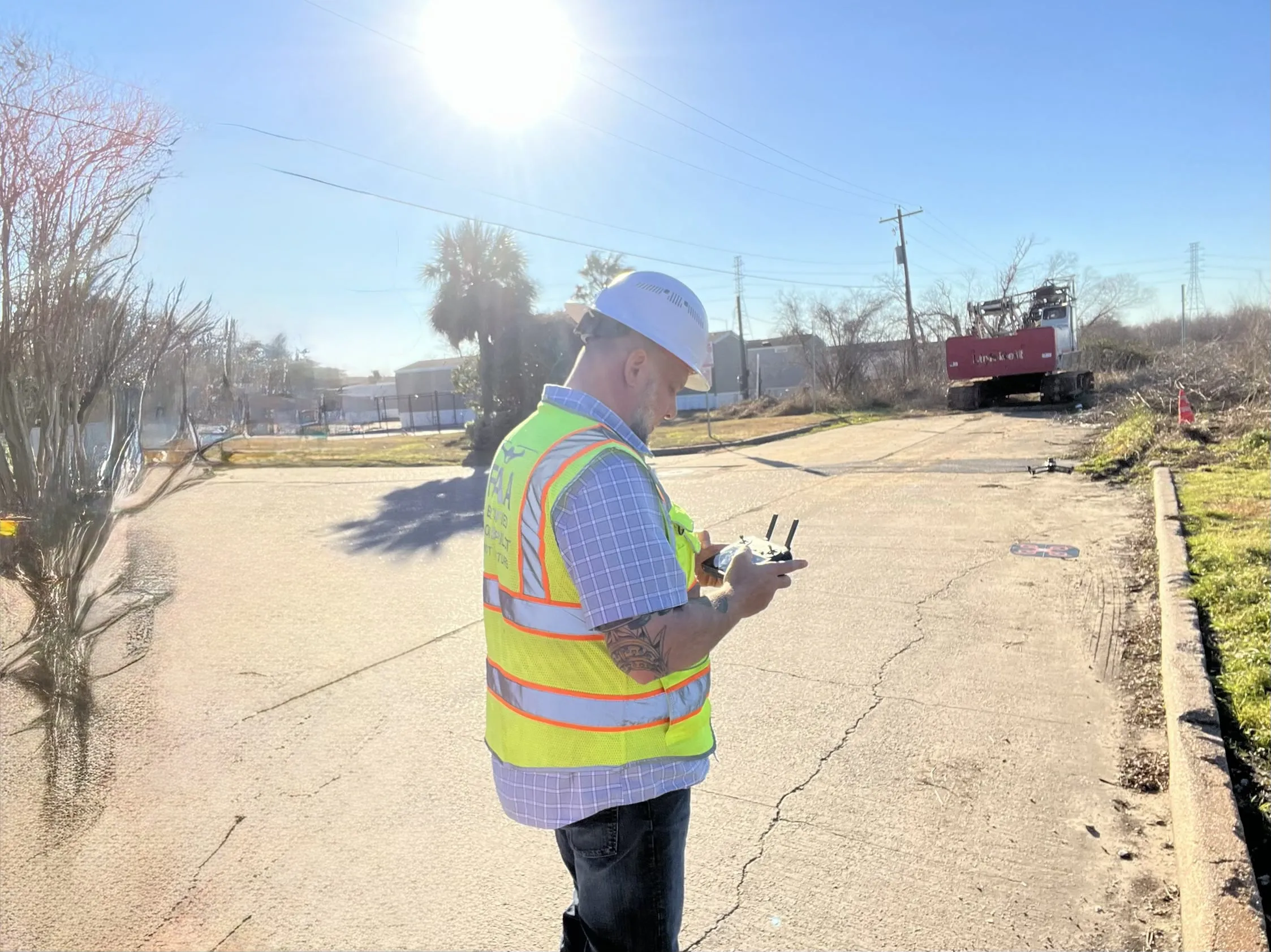 Axis pilot in a hard hat and high-visibility vest at the controller on a cleared industrial lot, tracked equipment working in the background.